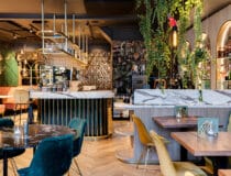 Kleurrijk restaurant met een verfijnde mamerlook op de tafelbladen, aan de bar en op de bartafels door decoratief plaatmateriaal FB38 Talco van Cleaf, welke een uitstraling van Carrara marmer in wit met donkergrijze aders heeft.